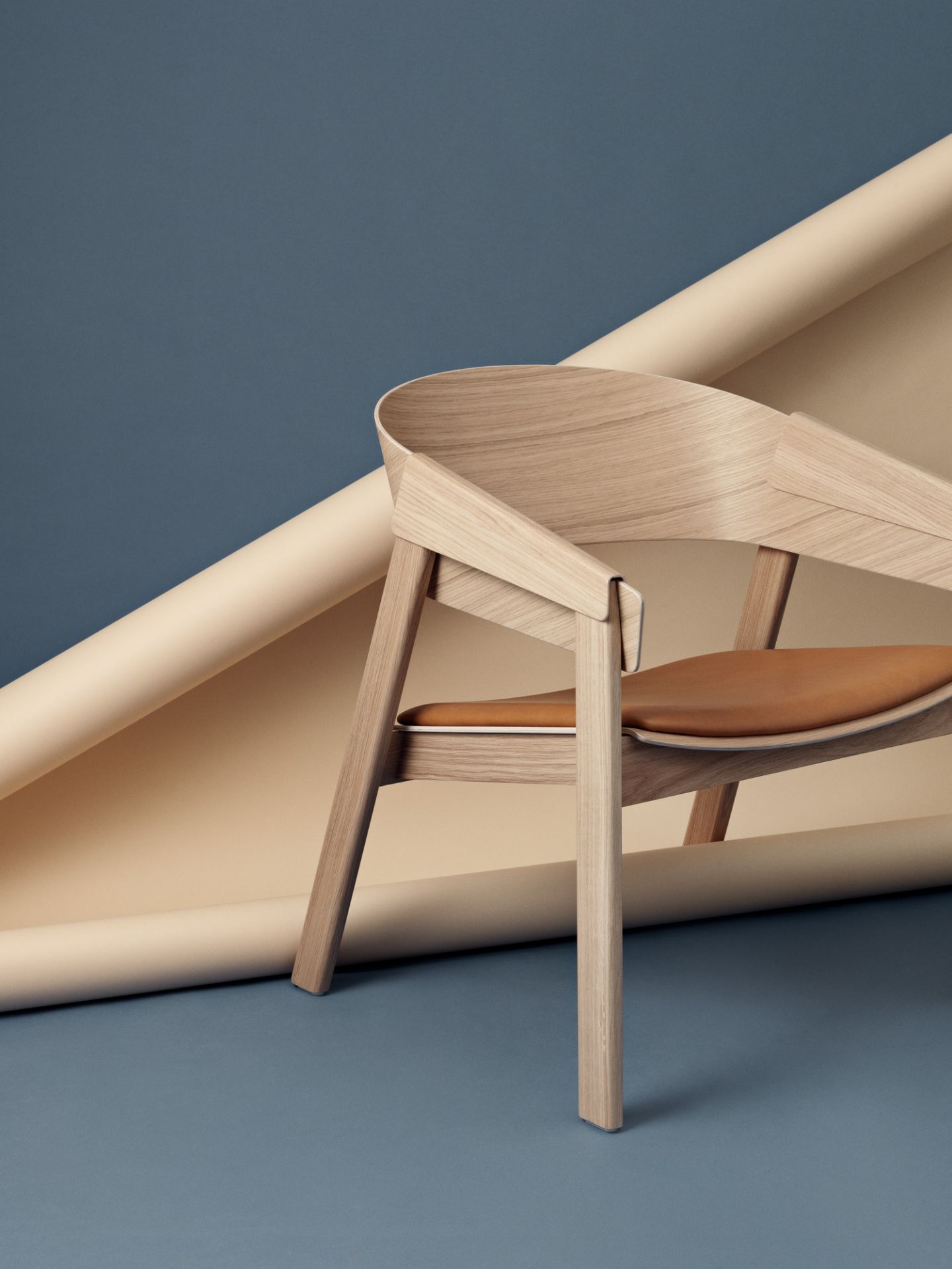 Muuto Cover Lounge Chair Stuhl aus Eichenholz mit braunem Ledersitz vor blauem Hintergrund.