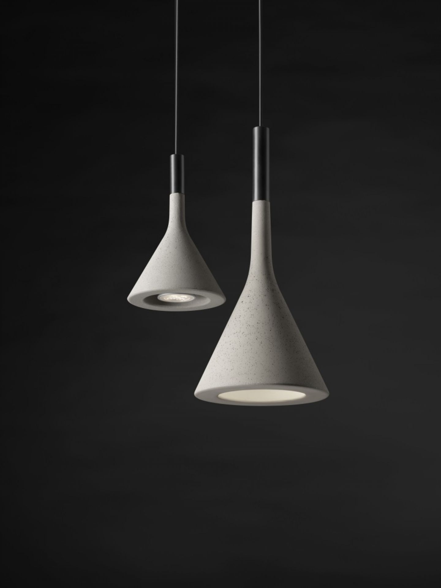 Zwei graue Aplomb Mini Pendelleuchten von Foscarini aus Beton vor dunklem Hintergrund.