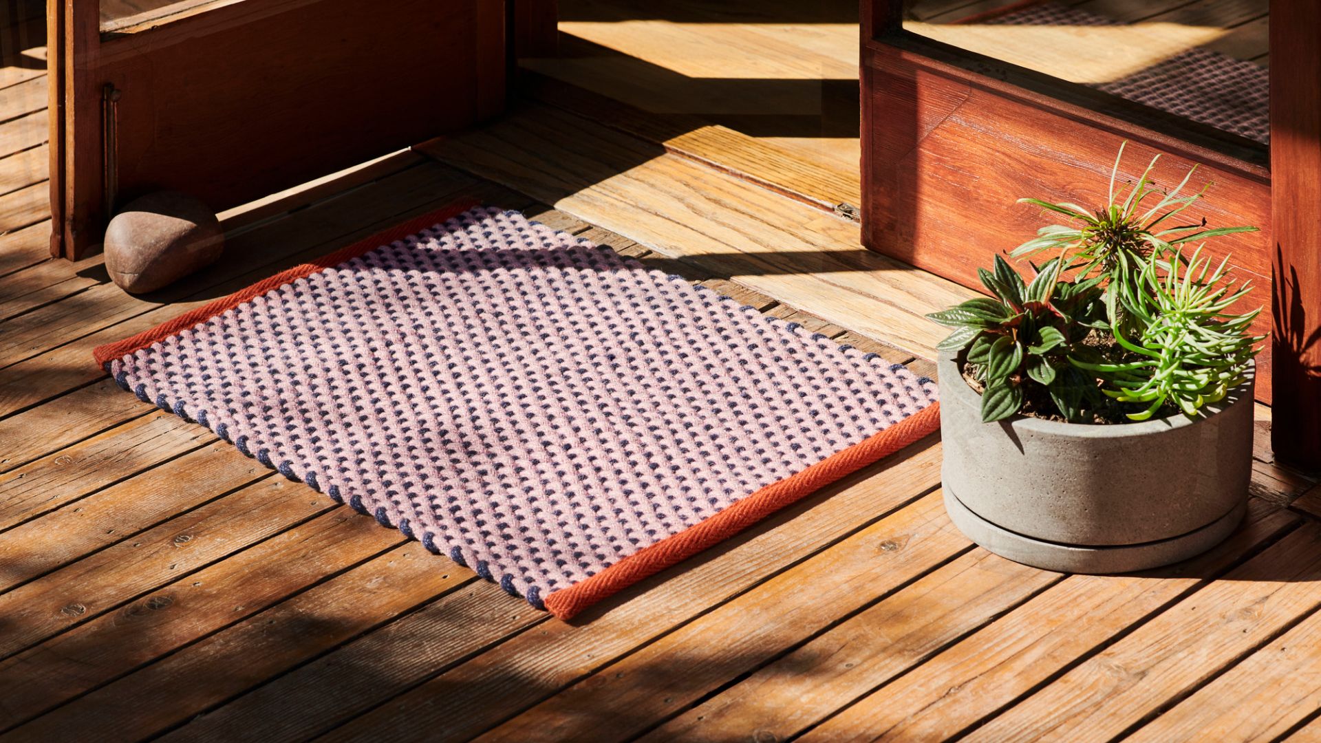 Hay Türvorleger Teppich auf Holzterrasse, rosa-blaues Webmuster mit orangefarbener Bordüre, neben Pflanztopf und offener Holztür.