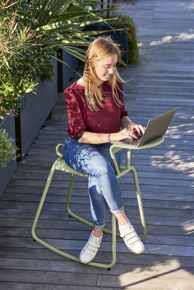 Frau arbeitet am Laptop auf Fermob Adadesk Beistelltisch Wippe Outdoor in Pistaziengrün auf Holzterrasse.
