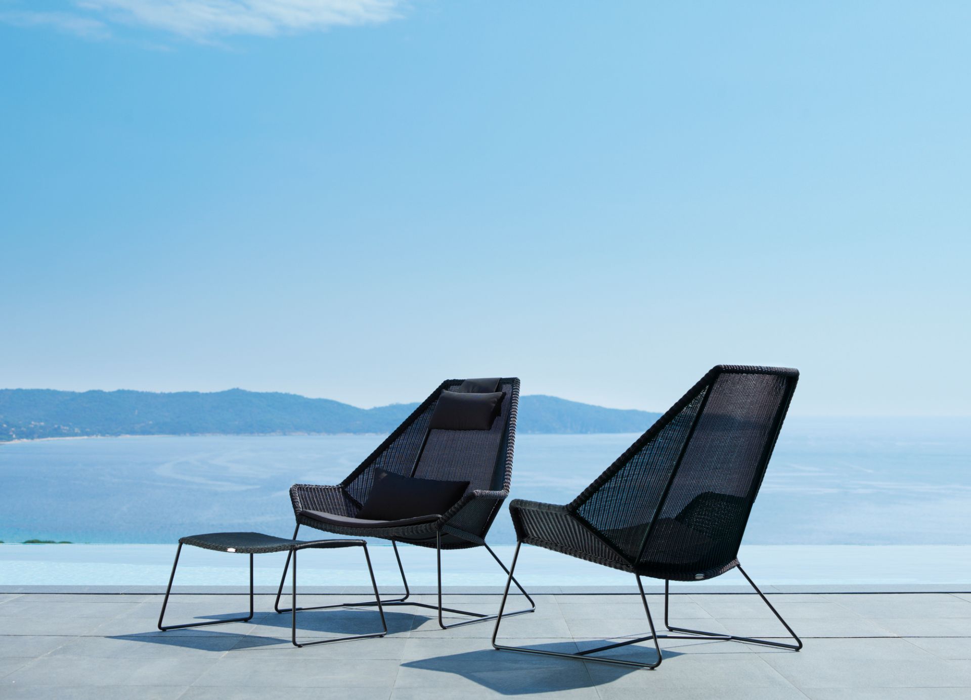 Zwei schwarze Breeze Outdoor Sessel von Cane-Line mit Hocker vor blauem Meer.