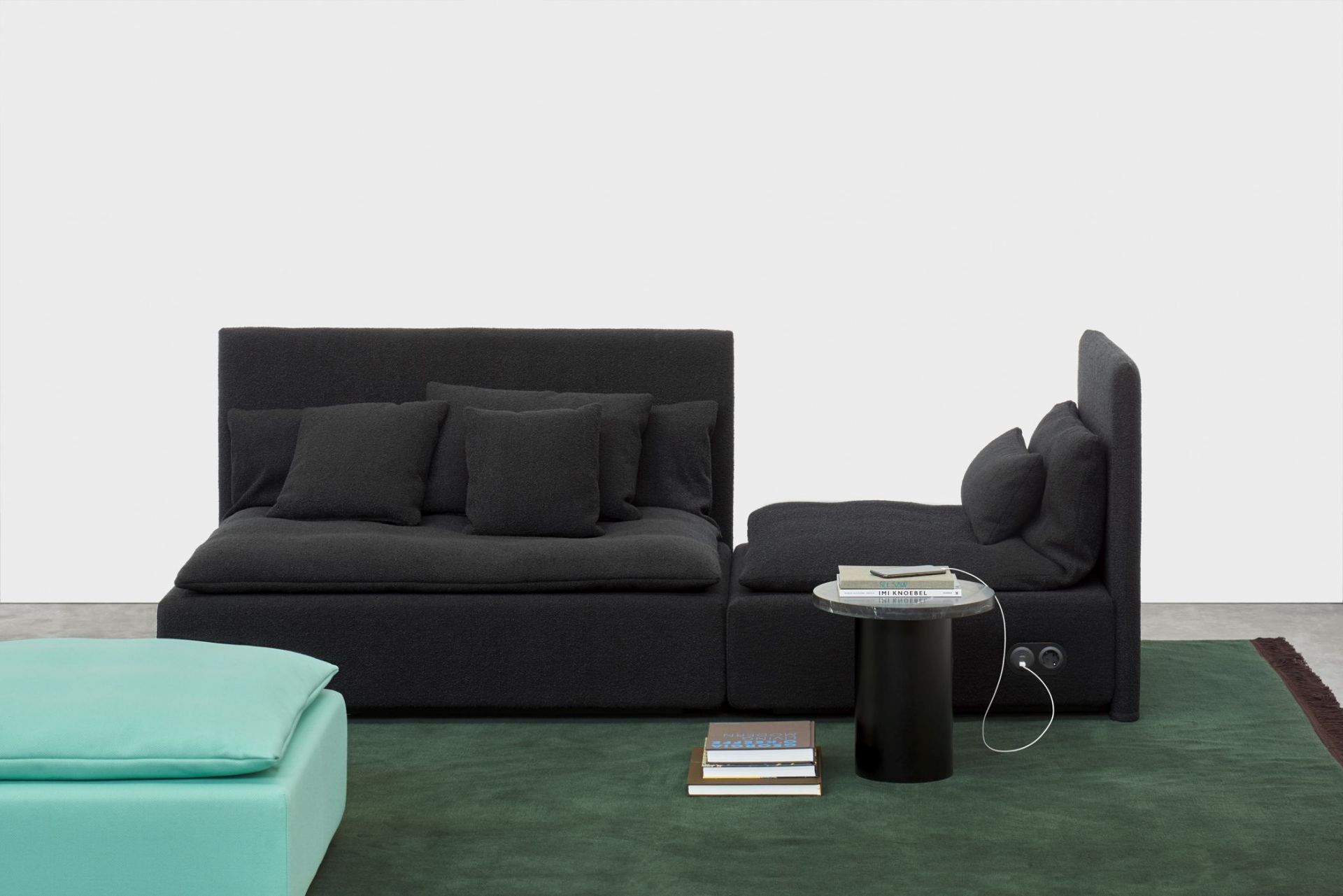 Enoki CT09 Beistelltisch von e15 neben modularem Sofa und türkisfarbenem Hocker auf grünem Teppich.