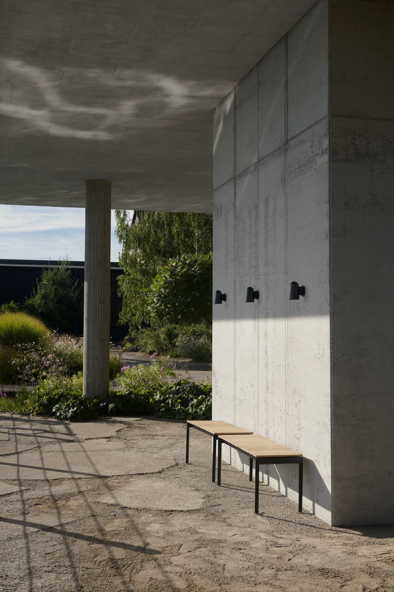 AndTradition Ville AV27 Outdoor Bank aus Holz und Metall vor Betonwand mit Außenleuchten.