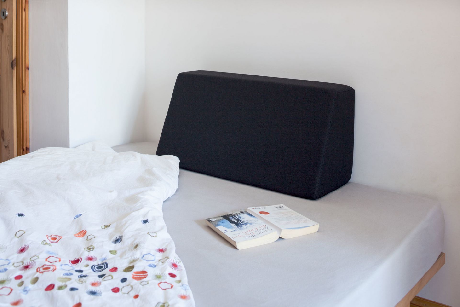 Schwarzes Tojo Lehn Rückenkissen auf einem Bett mit weißer Bettwäsche und Buch.