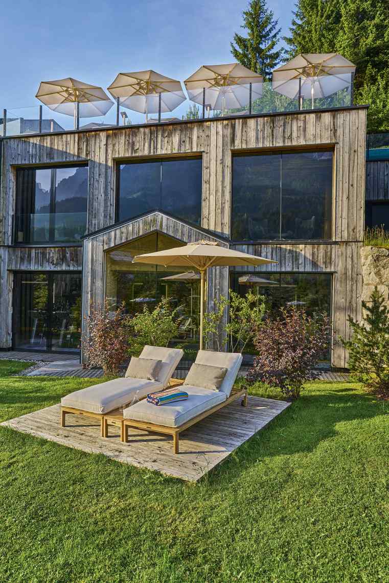 Beiger Weishäupl Schirm spendet Schatten über zwei Liegestühlen im Garten eines modernen Holzhauses.