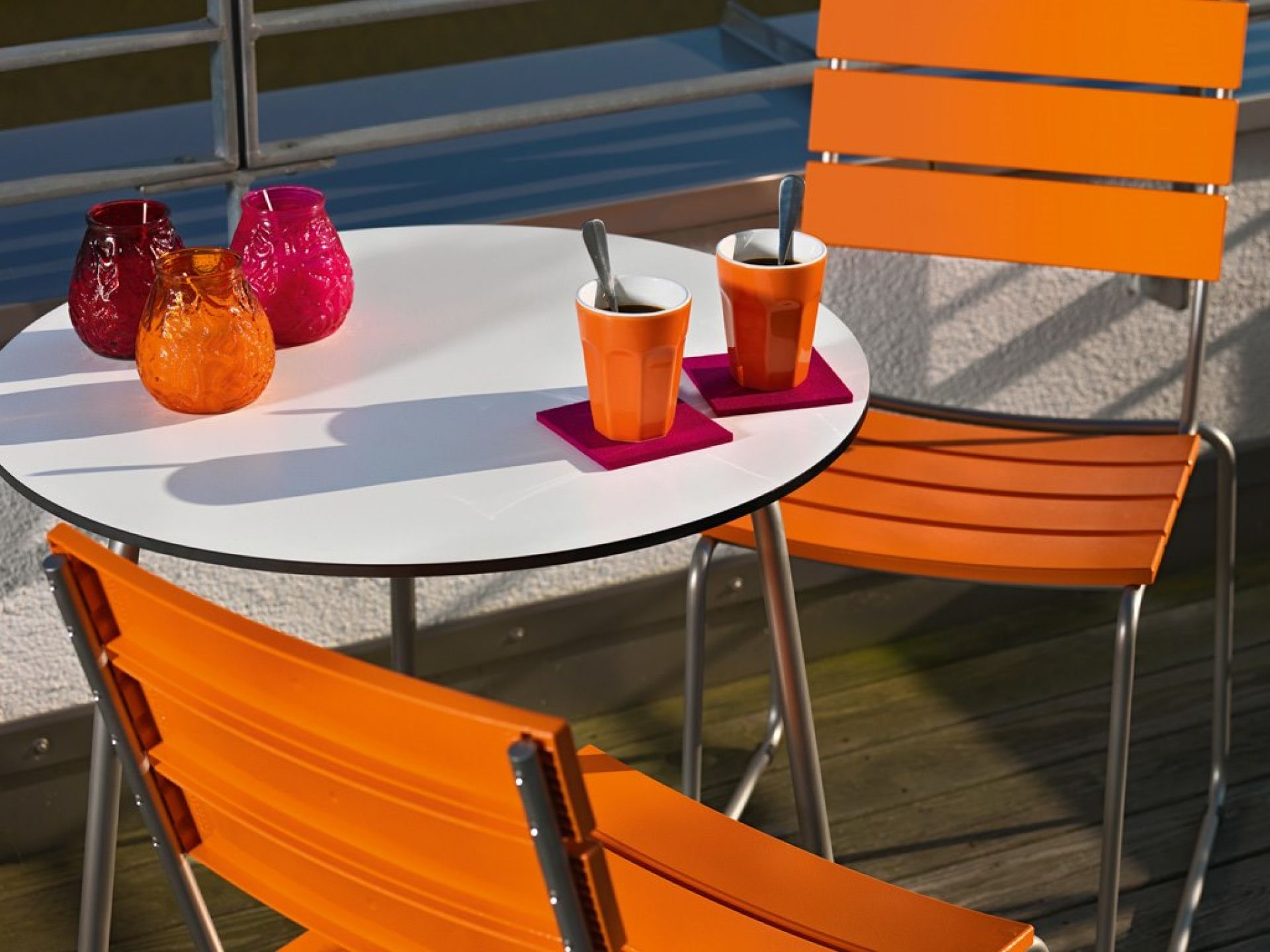 Weishäupl Bistrotisch mit Edelstahlgestell, orangefarbene Stühle, bunte Windlichter und Kaffeetassen auf einem Balkon.