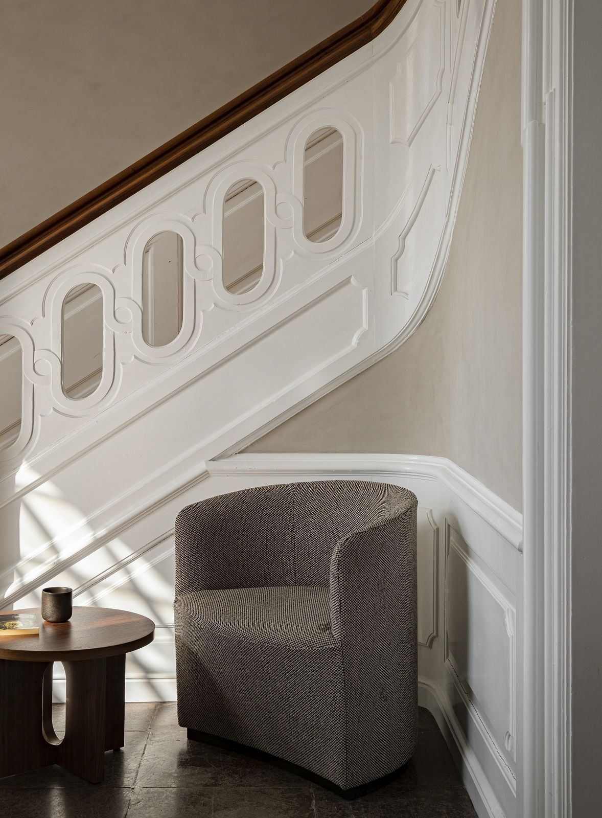 Tearoom Lounge Chair Sessel von Audo Copenhagen in grau-braunem Stoffbezug neben Treppe und Beistelltisch.