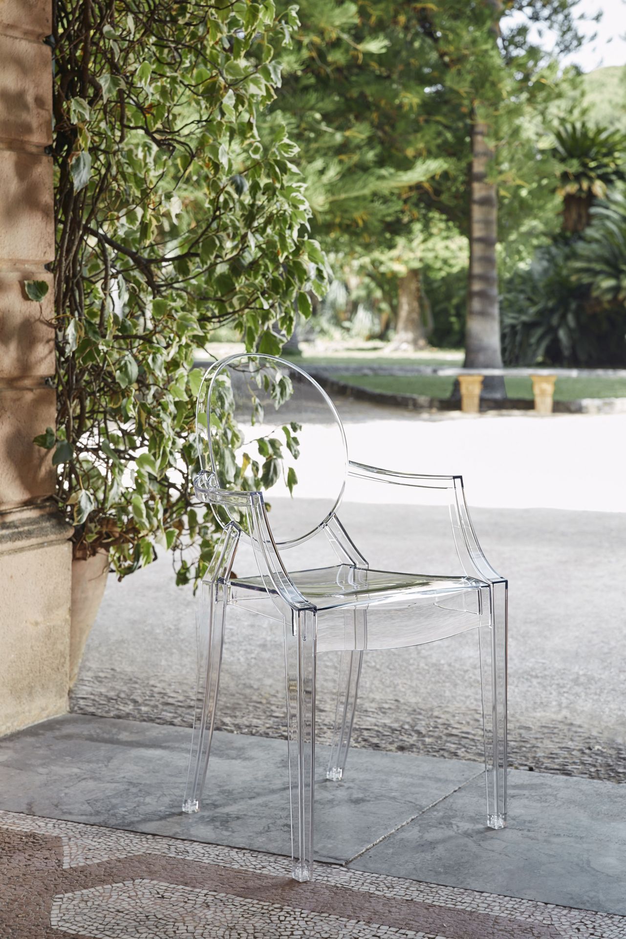 Glasklarer Louis Ghost Armlehnenstuhl von Kartell vor Grünpflanzen im Außenbereich. Designstuhl aus transparentem Kunststoff.