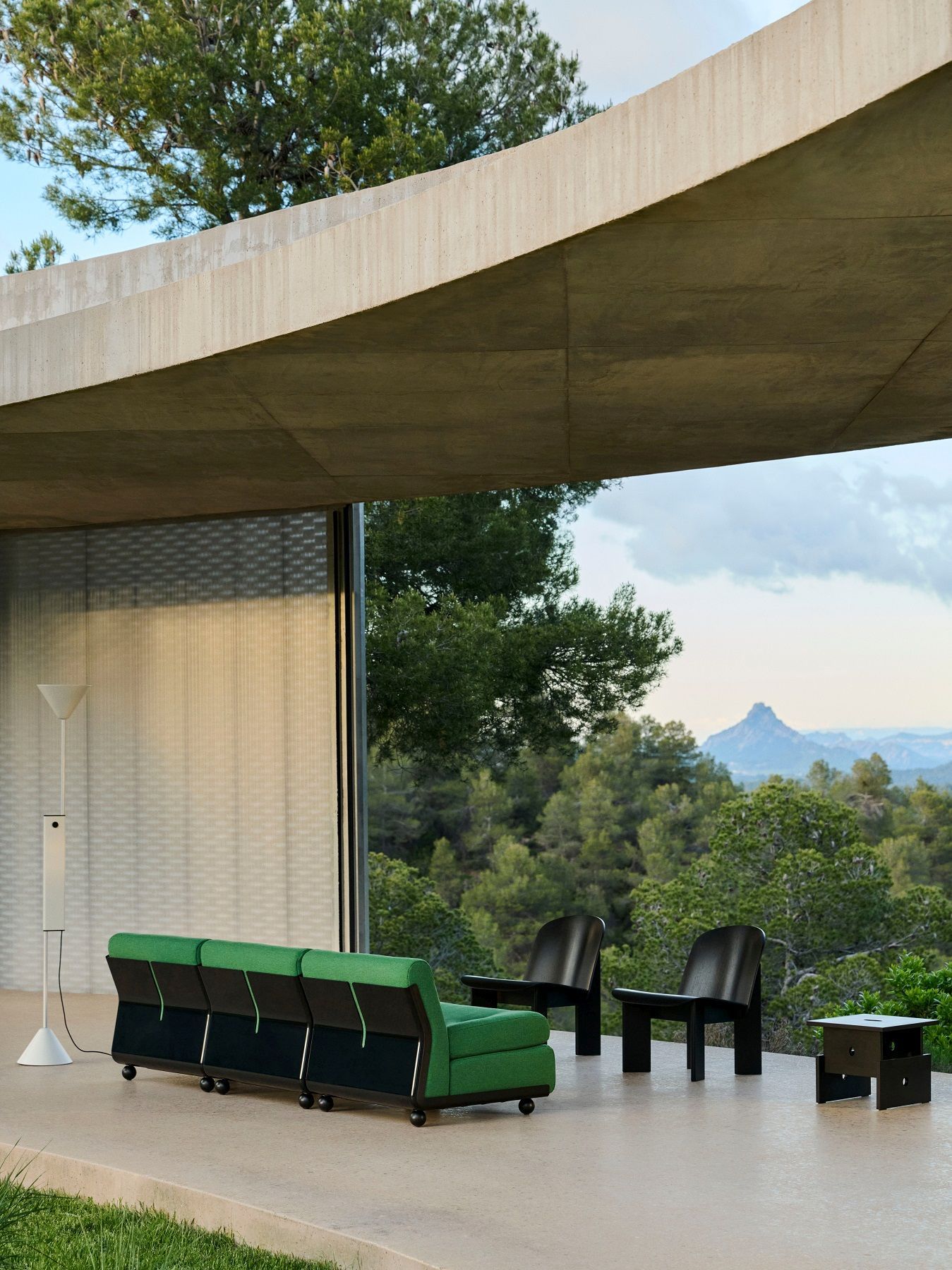 Weiße Twist Floor Lamp Stehleuchte von Hay neben grüner Couch und schwarzen Stühlen auf Terrasse mit Ausblick.