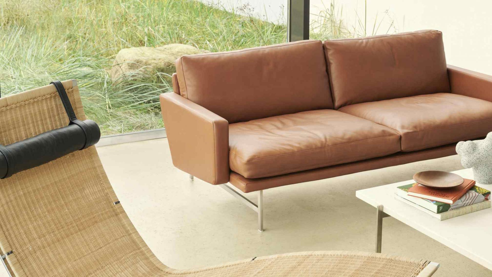 PK 80 Liege von Fritz Hansen, braunes Ledersofa und Rattansessel in modernem Wohnzimmer mit Tageslicht.