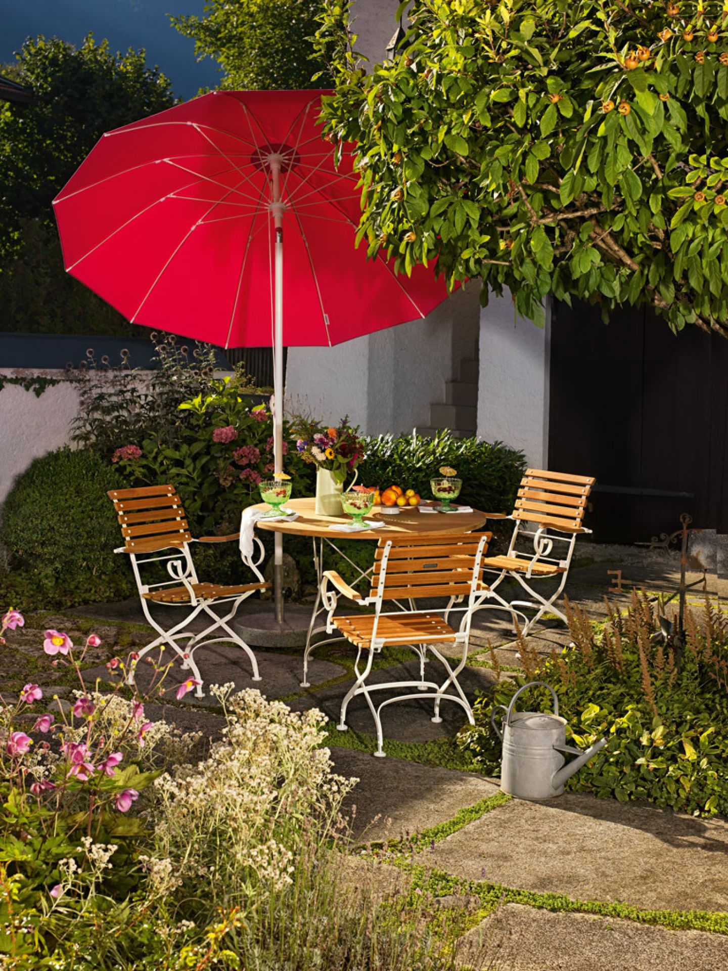 Weishäupl Classic Table klappbarer Gartentisch Ø110 cm, weiß, mit Stühlen und rotem Sonnenschirm im Garten.