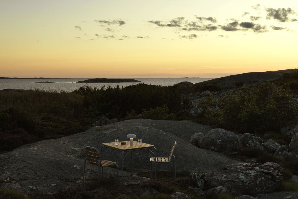 Normann Copenhagen Vig Tisch 90x90 cm Outdoor mit Stühlen und Tischleuchte vor Küstenlandschaft.