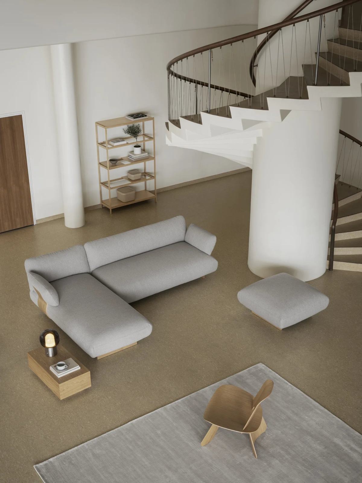 Luftaufnahme eines hellen Wohnzimmers mit grauem Ecksofa, Hocker und Blomus Nagomi Holzstuhl auf Teppich.