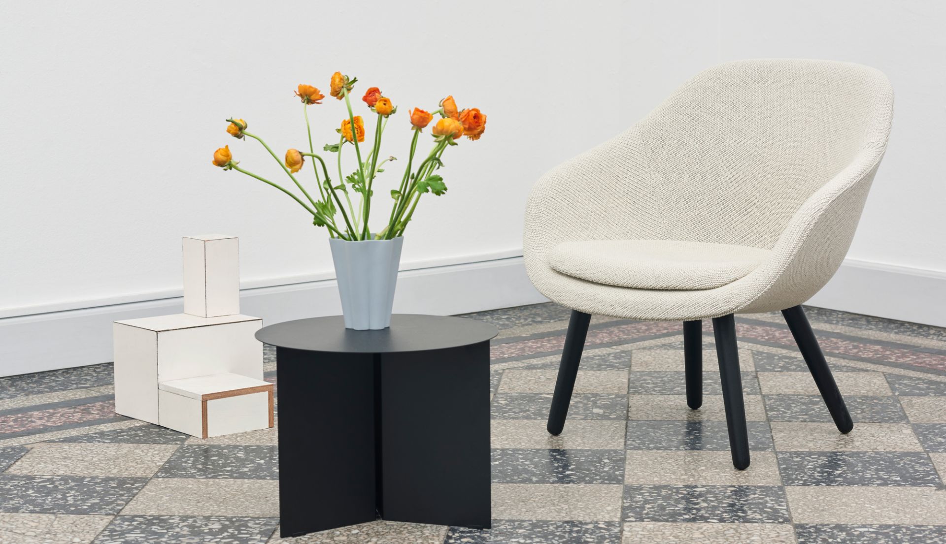 Beiger About a Lounge AAL 82 Sessel von Hay neben schwarzem Tisch mit Blumen auf Schachbrettboden.