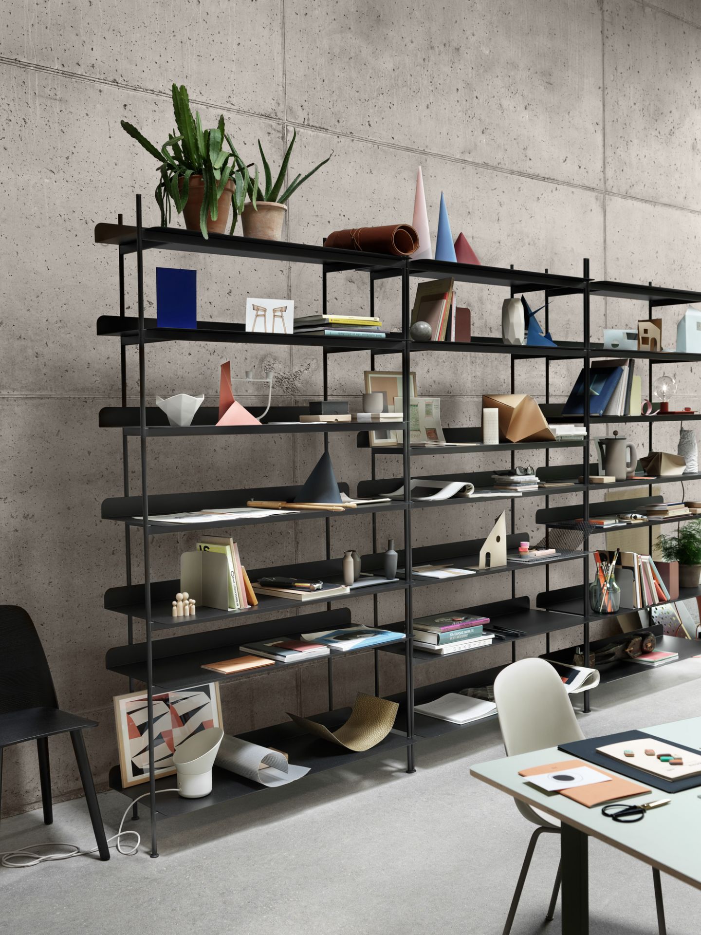 Schwarzes, modernes Regalsystem vor einer Betonwand, dekoriert mit Büchern, Pflanzen und Büroaccessoires, im Vordergrund ein Muuto 70/70 Tisch.