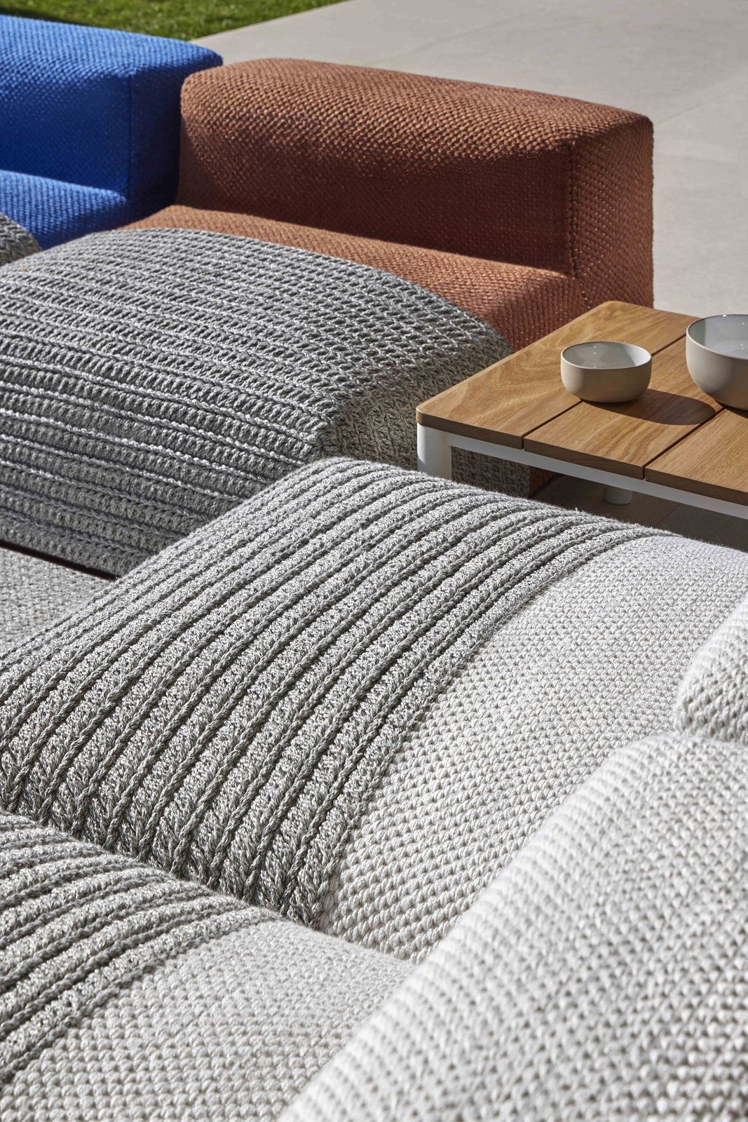 Detailaufnahme von grauen Mangas Outdoor Modulen, einem braunen und einem blauen Outdoor Sessel und einem Holztisch.