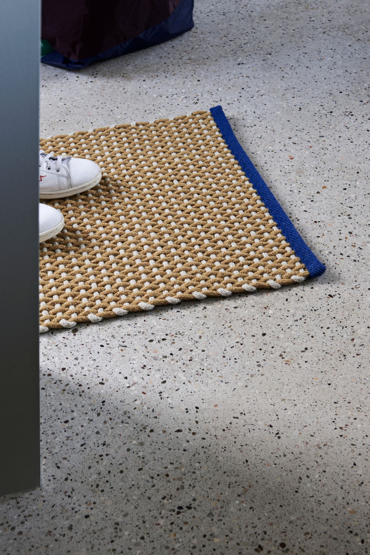 Beiger Hay Teppich mit blauem Rand und weißen Schuhen auf grauem Boden. Fußmatte für den Eingangsbereich.