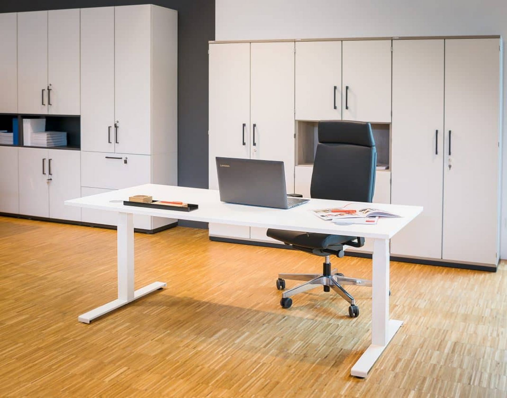 Weißer Hiplift Sitz-Stehtisch mit Laptop und Bürostuhl vor weißen Büroschränken. Elektrisch höhenverstellbarer Schreibtisch.