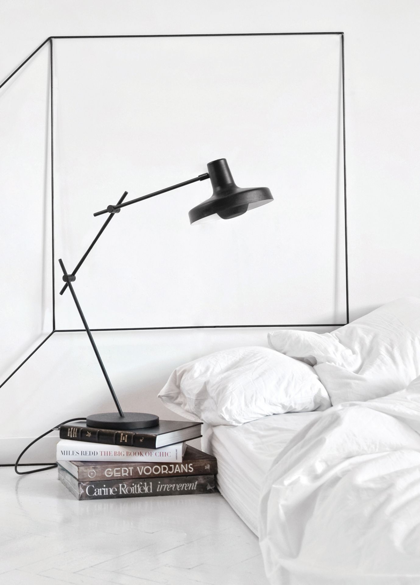 Arigato Tischleuchte von Grupa, schwarze Designlampe auf Bücherstapel neben einem weißen Bett.