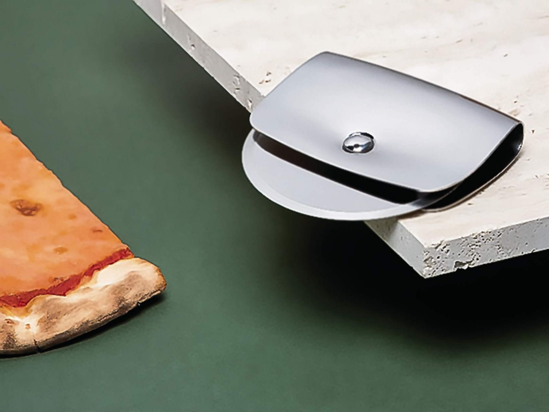 Alessi VS04 Taio Pizzaschneider aus Edelstahl neben einem Stück Pizza auf grünem Hintergrund.