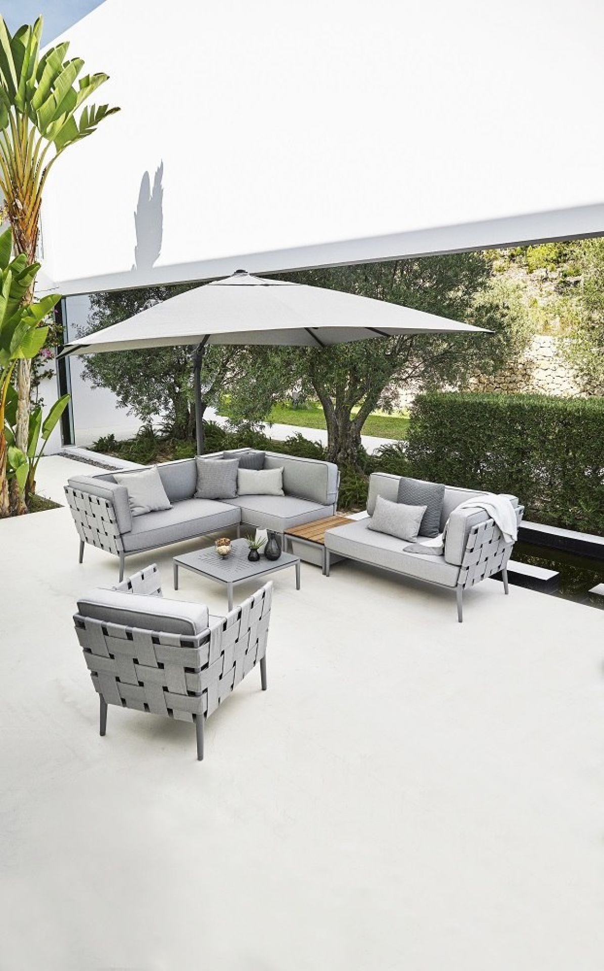 Weißer Hyde Luxe Sonnenschirm von Cane-Line mit Fuß über grauer Lounge-Garnitur im Garten.