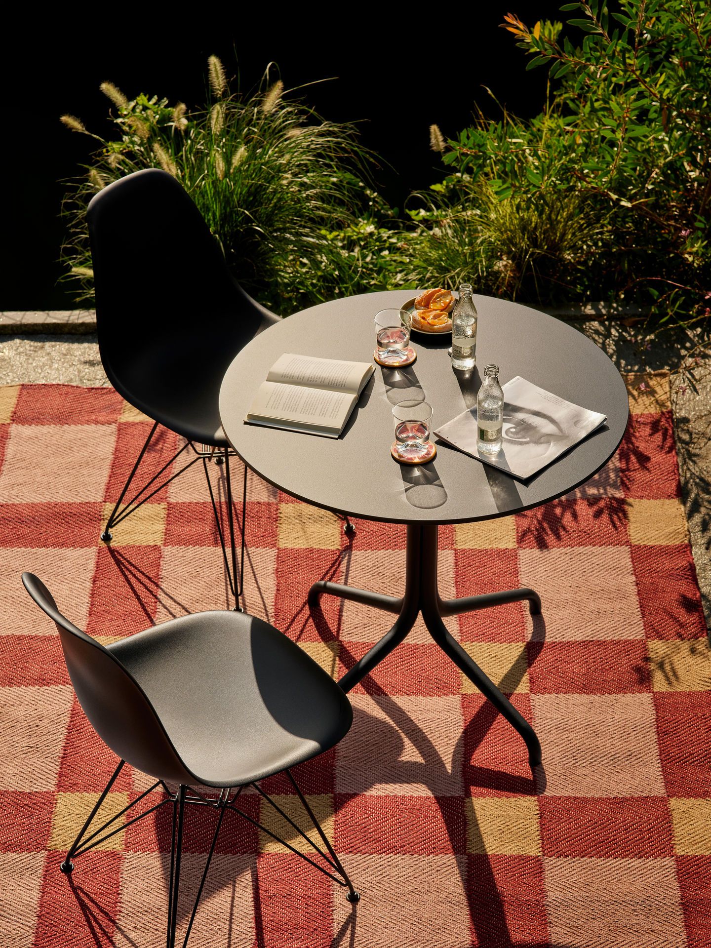 Zwei Eames Stühle und ein runder Tisch auf einem rot-gelb karierten Teppich im Außenbereich.