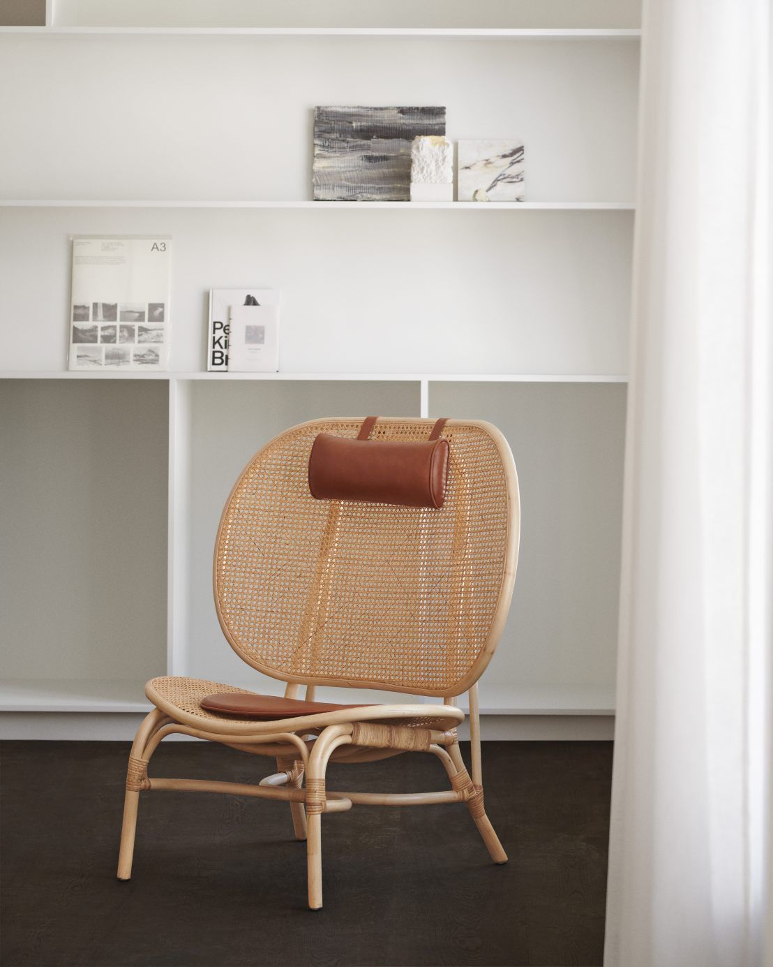 NORR11 Nomad Chair Sessel aus Rattan mit braunem Lederkissen vor weißem Regal.