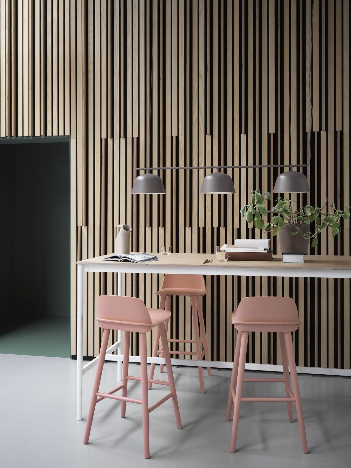 Graue Muuto Rail Lamp Pendelleuchte über Esstisch mit rosa Barhockern und Holzpaneelen.