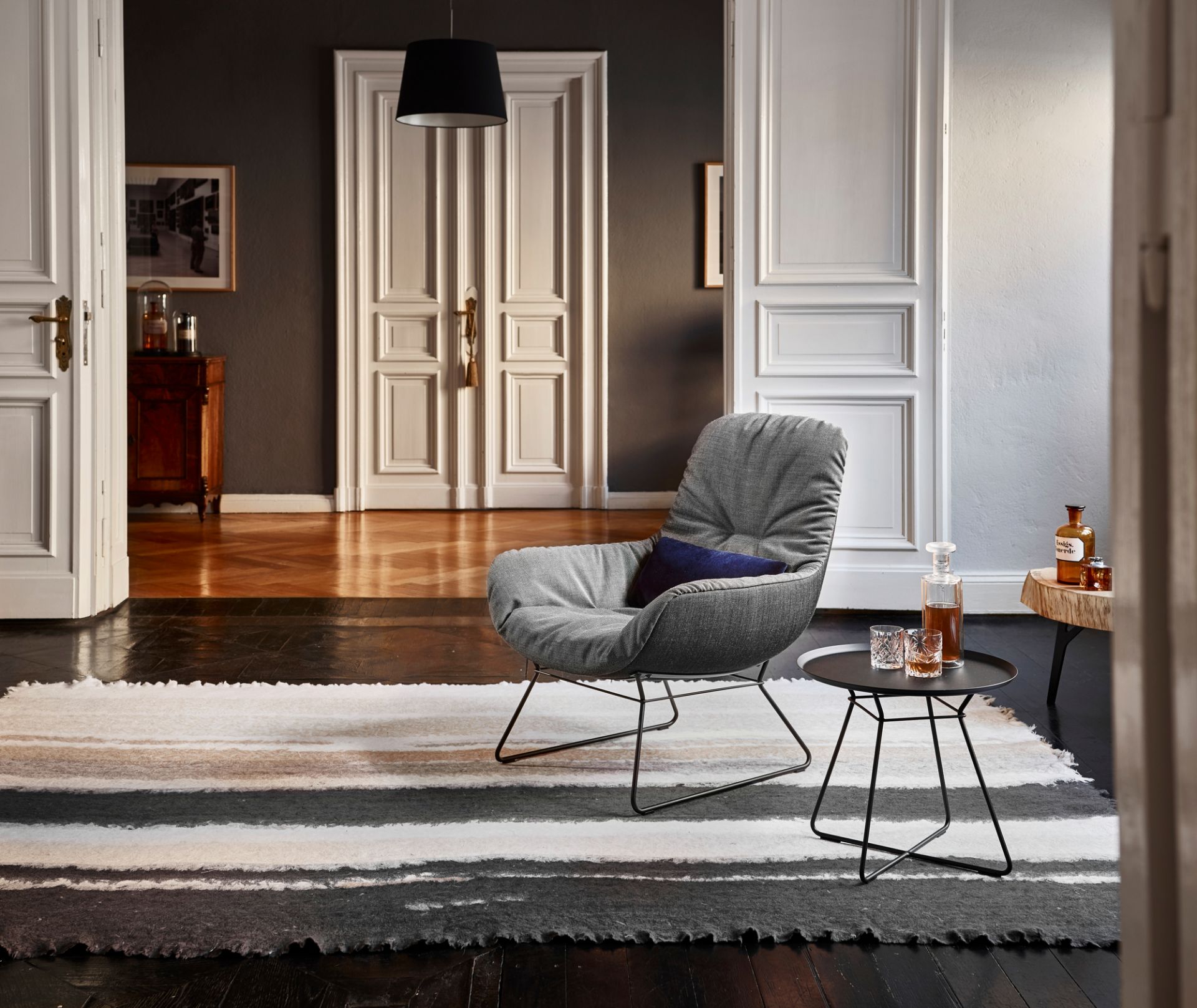 Grauer Leya Cocktail Loungesessel Freifrau Manufaktur mit Kissen und Beistelltisch in stilvollem Interieur.