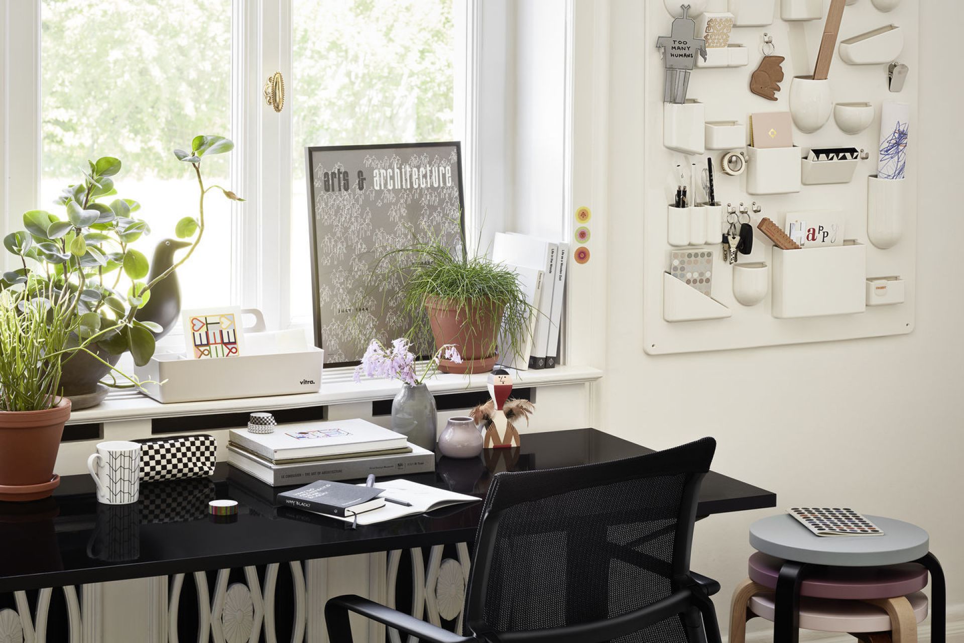 Detailaufnahme eines Schreibtisches mit Vitra Holzfigur, Pflanzen, Büchern und Büro-Accessoires vor einem Fenster.