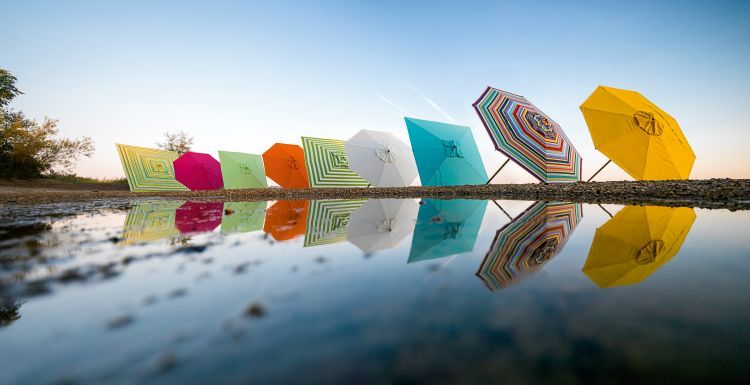 Mehrfarbige Weishäupl Sonnenschirme am Strand, reflektiert im Wasser. Klassischer Sonnenschutz mit Flaschenzug und Knickgelenk.