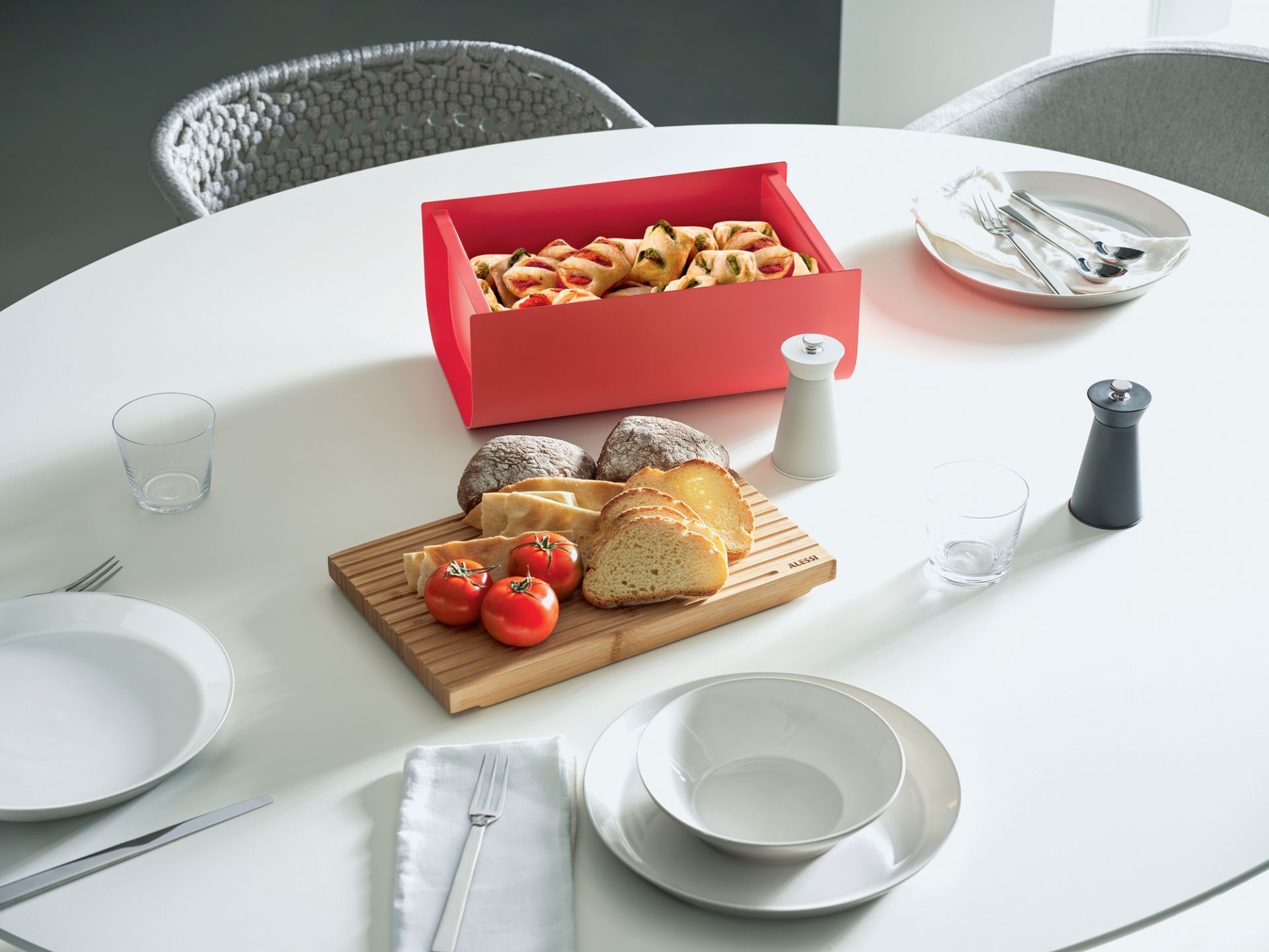 Roter Mattina Brotkasten von Alessi auf einem gedeckten Tisch mit Brot, Tomaten und Gebäck.