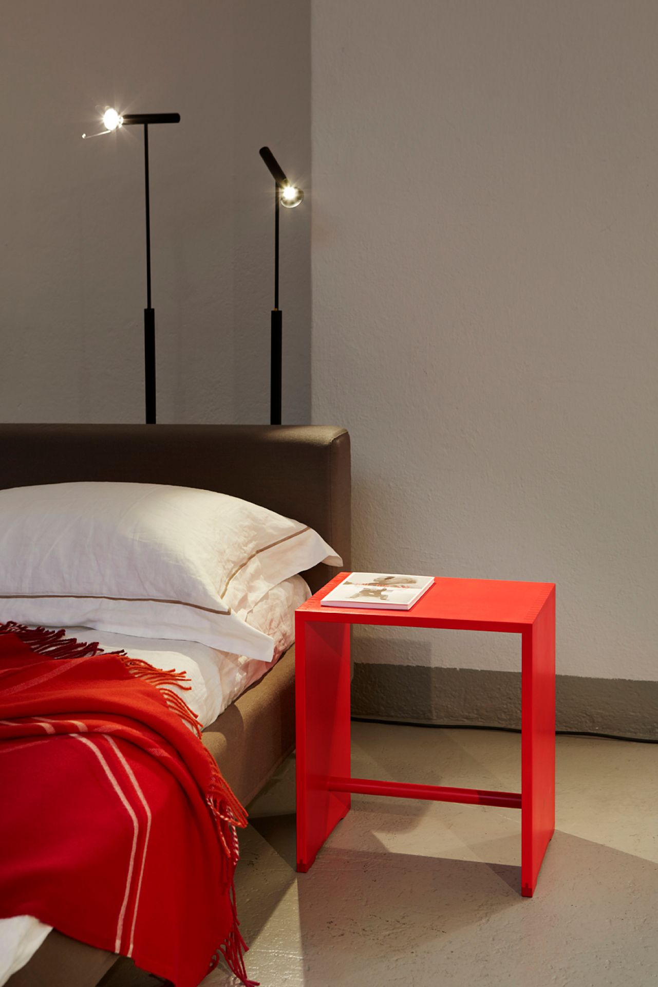 Roter Ulmer Hocker Max Bill als Nachttisch neben einem Bett mit roter Decke.