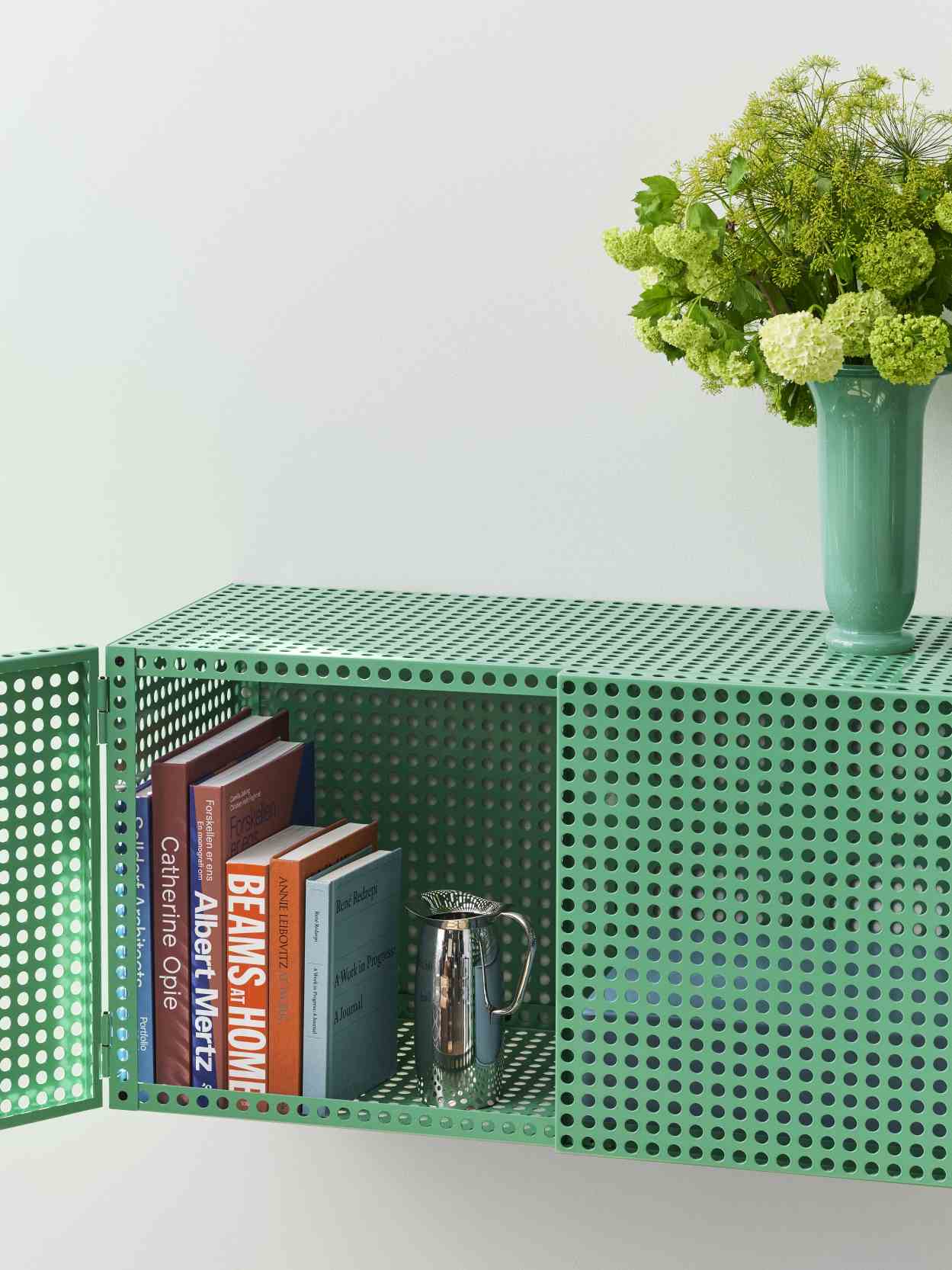 Grünes Hay Perforated Cabinet Wall Sideboard mit Büchern, einer Kanne und einer Vase mit Blumen.
