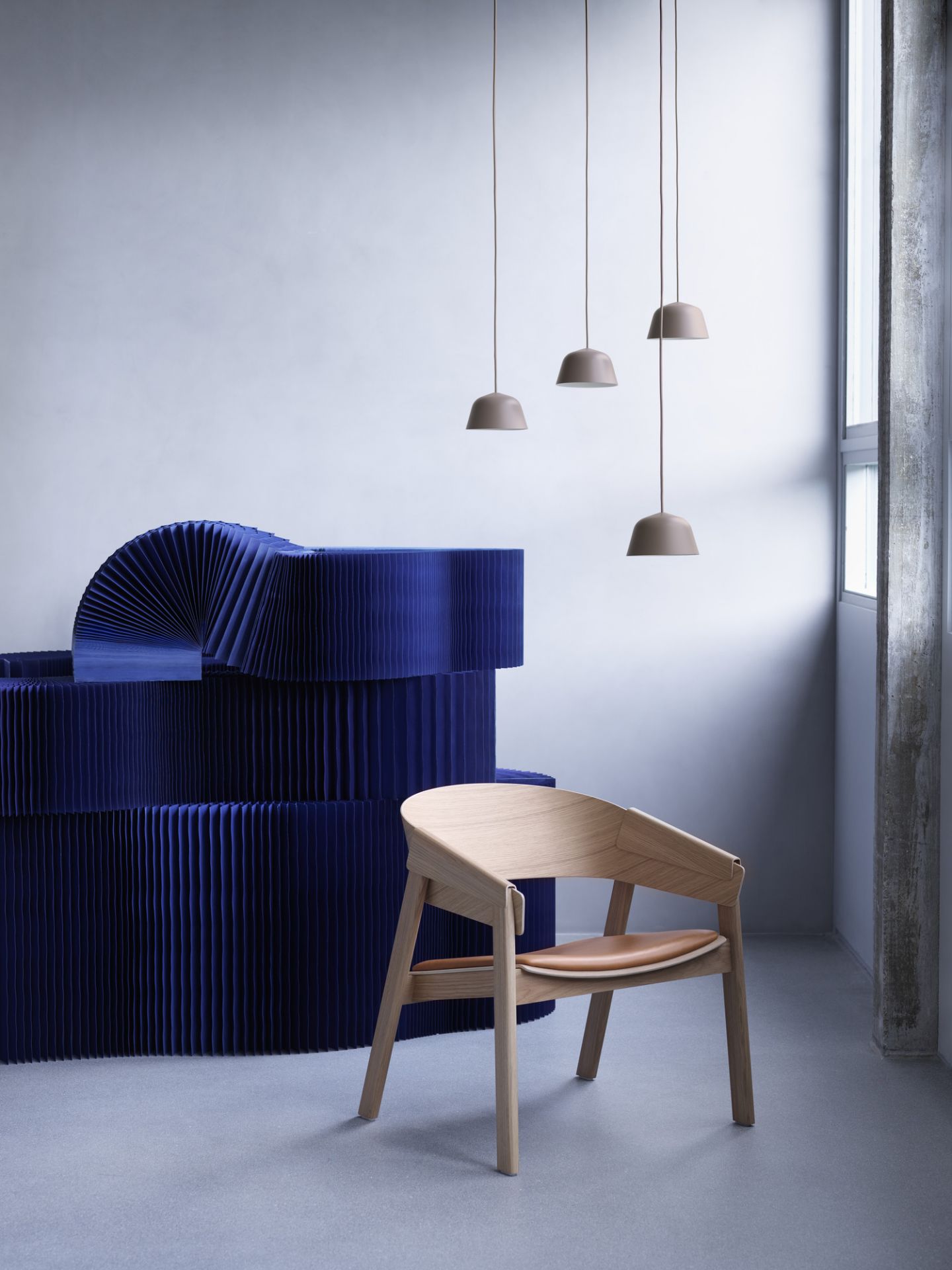 Muuto Cover Lounge Chair Stuhl aus hellem Holz mit braunem Ledersitz vor blauer Kulisse.