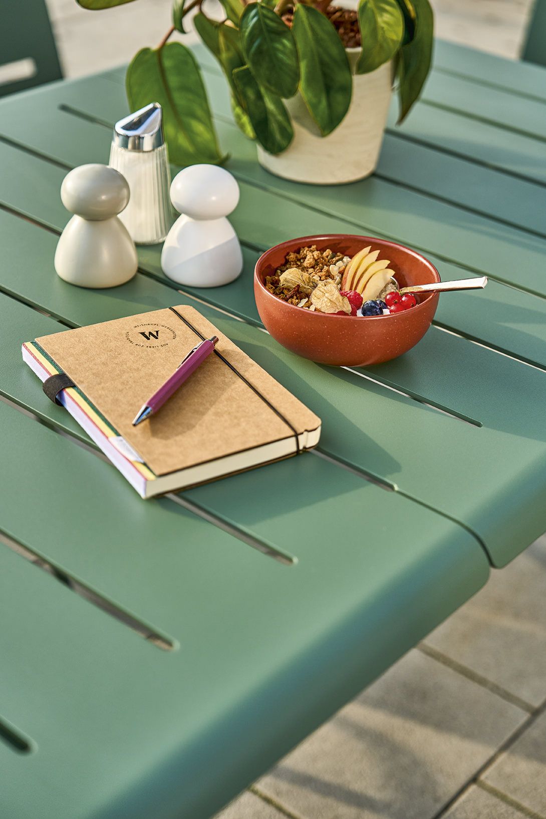 Vapio Tisch Outdoor von Weishäupl, Gartentisch in Grün mit Frühstücks-Szene, Müsli, Notizbuch und Gewürzen.
