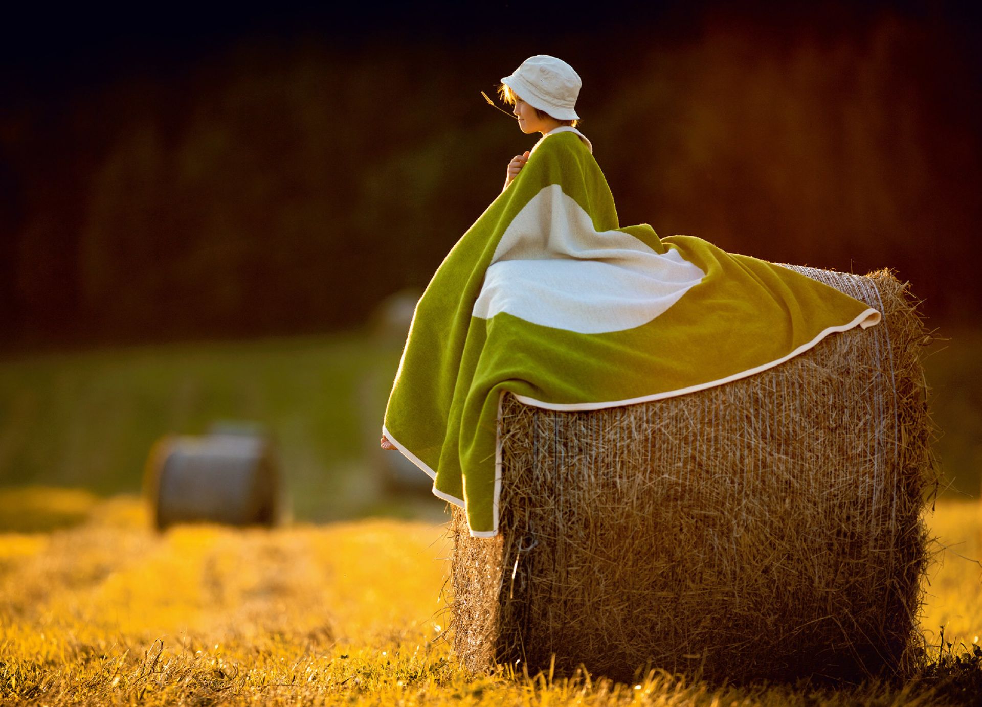 Kind sitzt auf Heuballen, gehüllt in grüne Vera Wolldecke von Pappelina, Kuscheldecke für gemütliche Stunden im Freien.