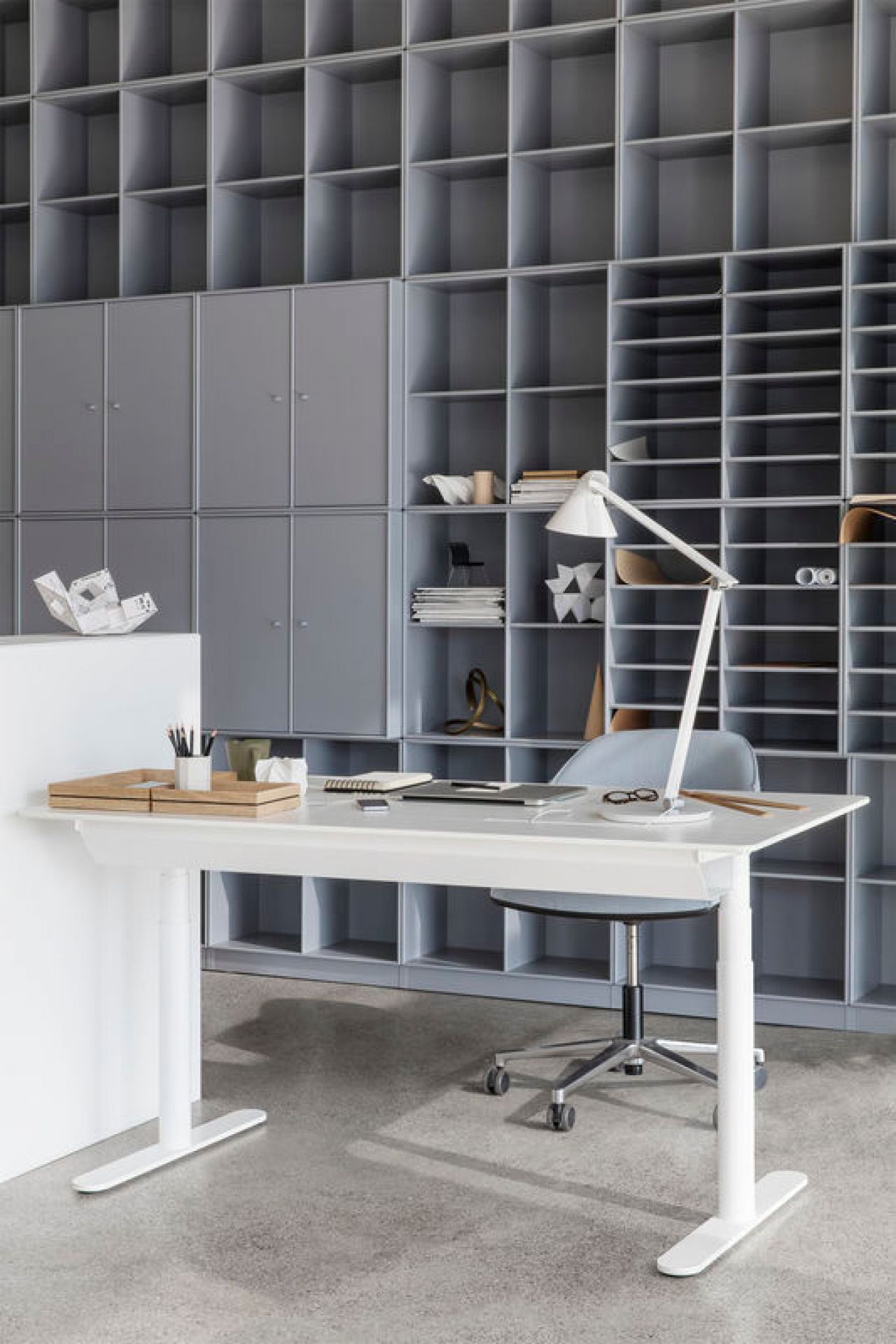 Weißer Hilow Tisch, höhenverstellbar, mit Schreibtischlampe, Laptop und Büromaterial vor grauem Regalsystem.