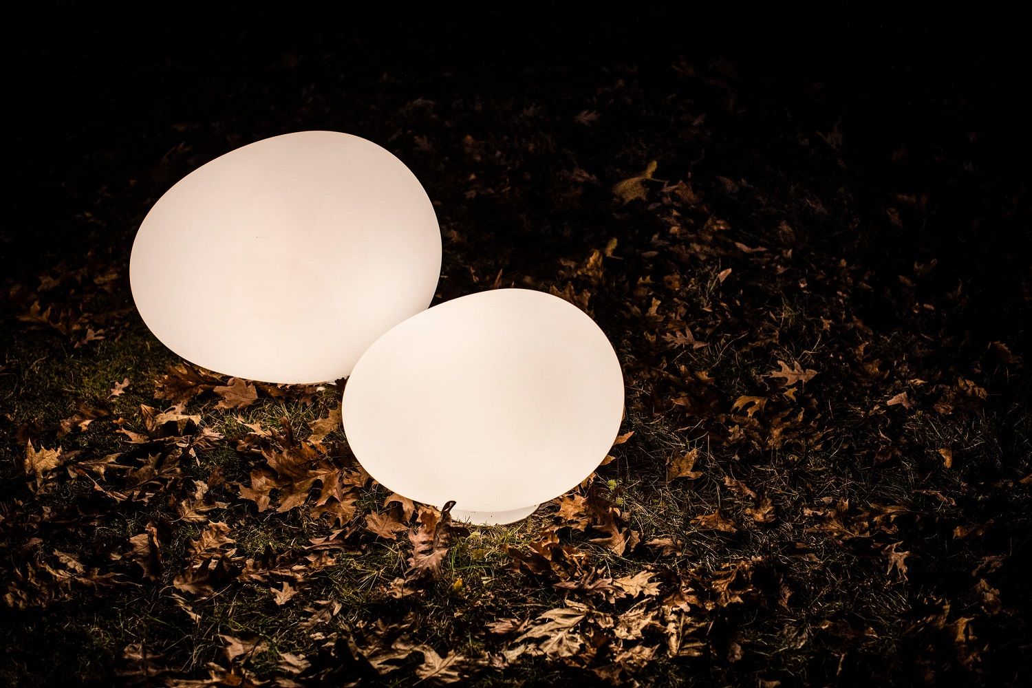 Zwei leuchtende Gregg Outdoor Bodenleuchten von Foscarini auf Rasen mit Herbstlaub im Dunkeln.