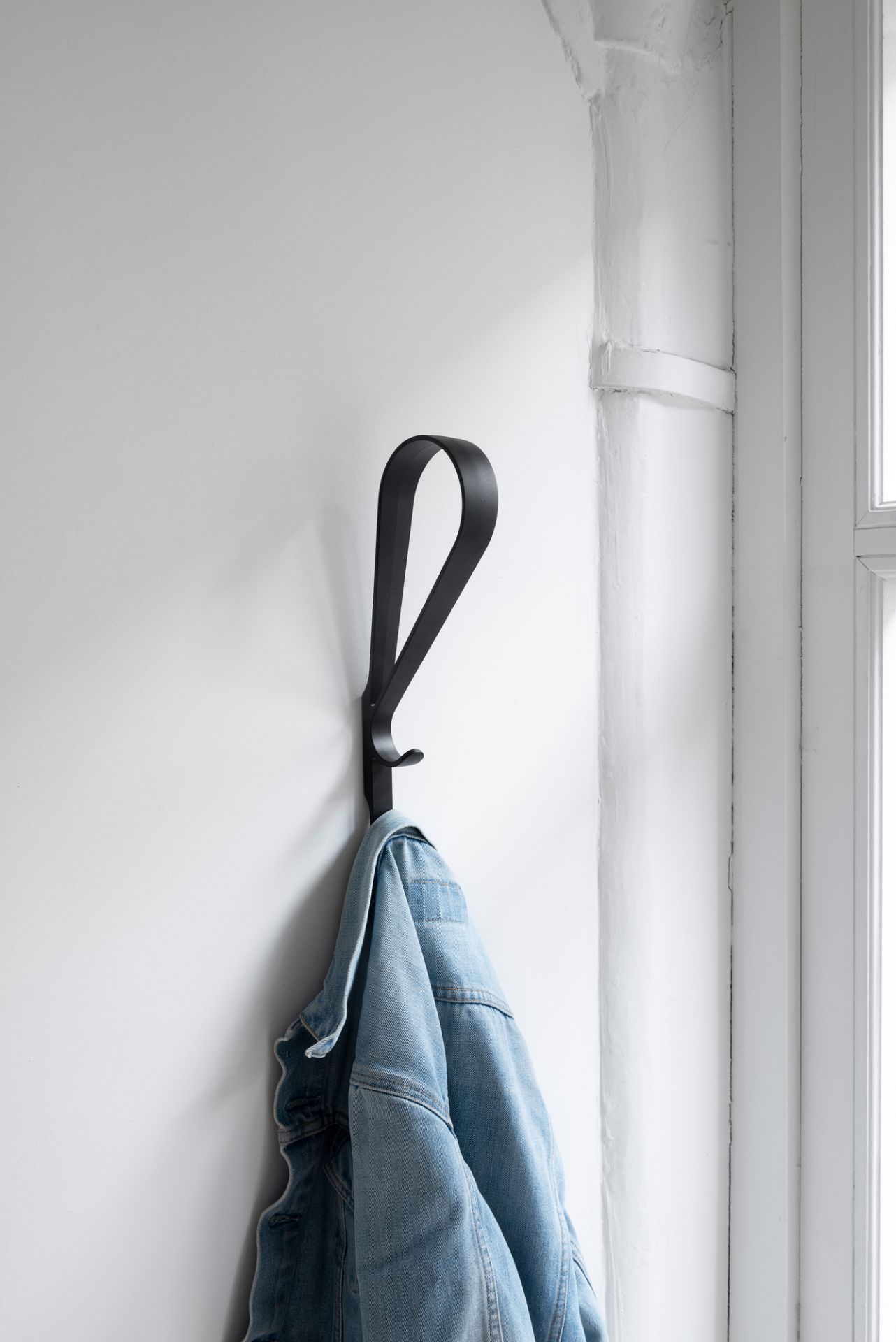 Schwarzer Artek Tupla Wandhaken an weißer Wand mit hängender Jeansjacke, moderne Garderobenlösung für Flur und Zuhause.