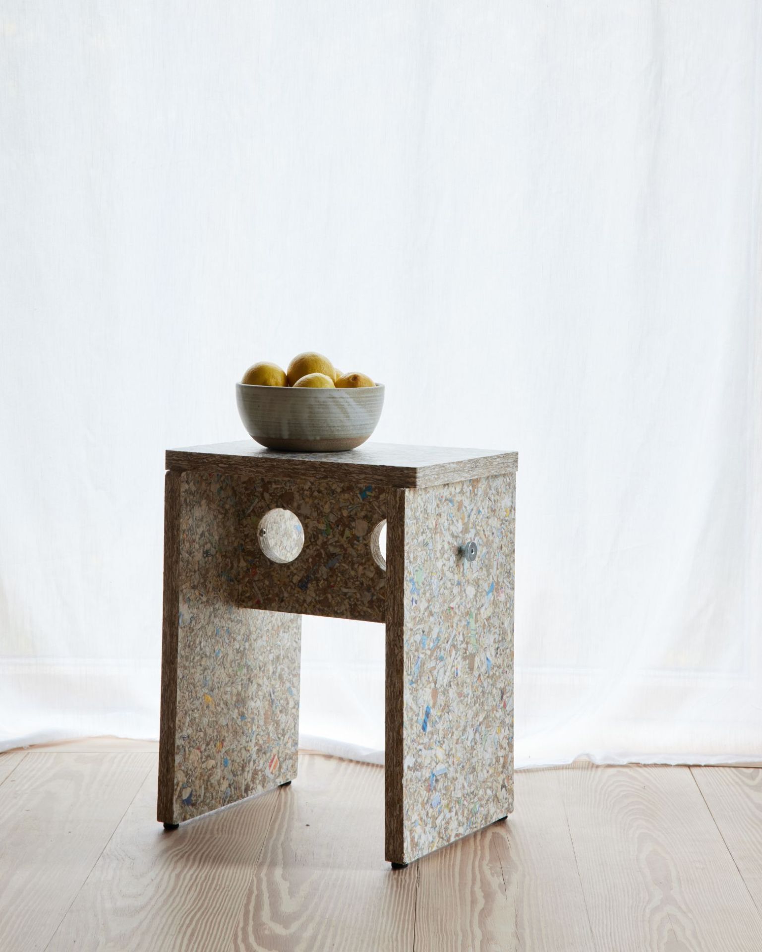 Remake Hocker aus recyceltem Material mit Schale Zitronen auf Holzboden vor weißem Vorhang.