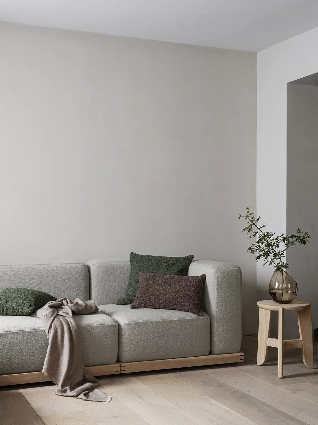 Heller Wohnraum mit Sofa, Kissen und einem Eli Stool Sitzhocker von Blomus mit Vase und Zweigen.