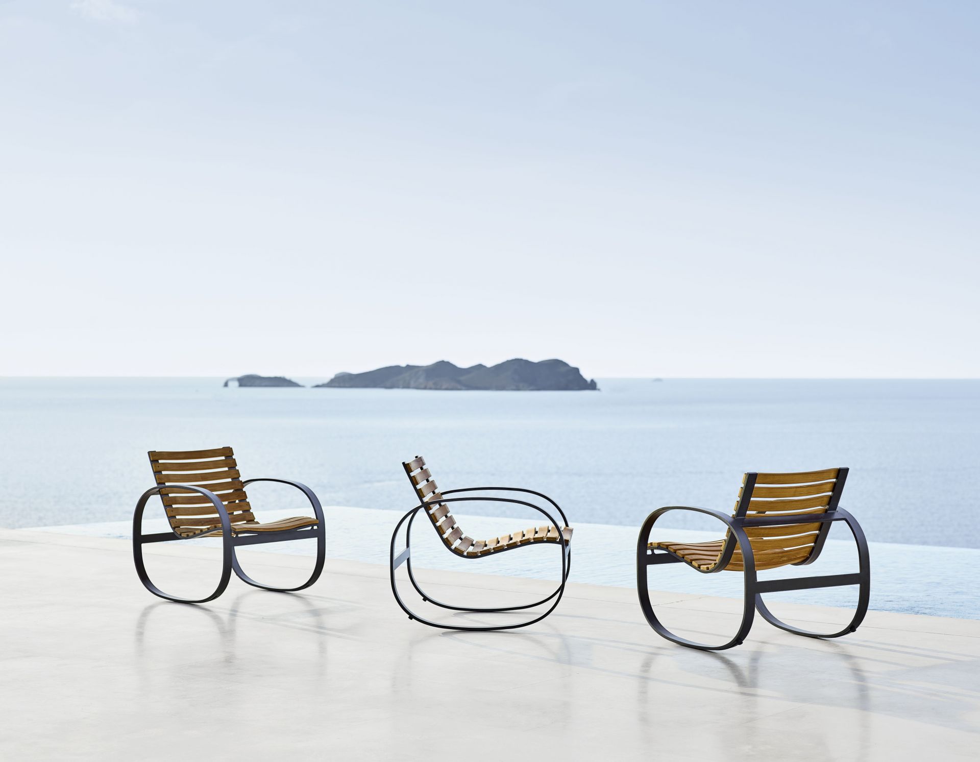 Drei Cane-Line Parc Schaukelstühle aus Teakholz und schwarzem Metall vor blauem Meer.