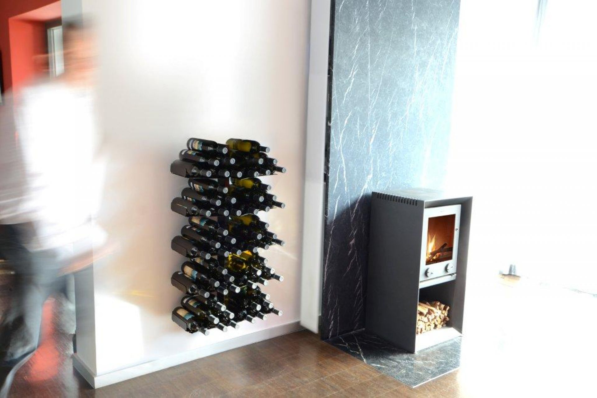 Weinregal "Wine Tree" an weißer Wand, gefüllt mit Weinflaschen, neben Kaminofen im modernen Wohnzimmer.