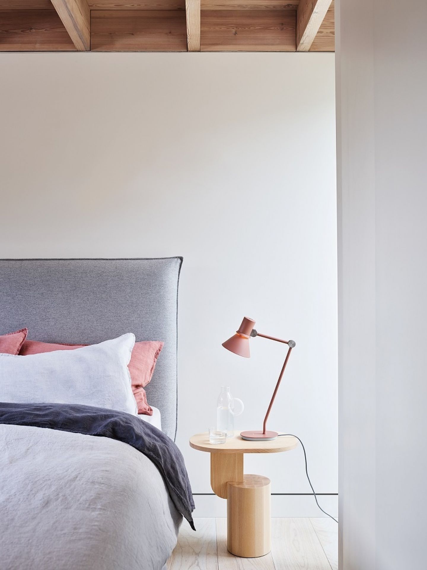 Rosa Anglepoise Type 80 Tischleuchte auf Nachttisch neben Bett mit grauer Bettwäsche und Holzbalkendecke.