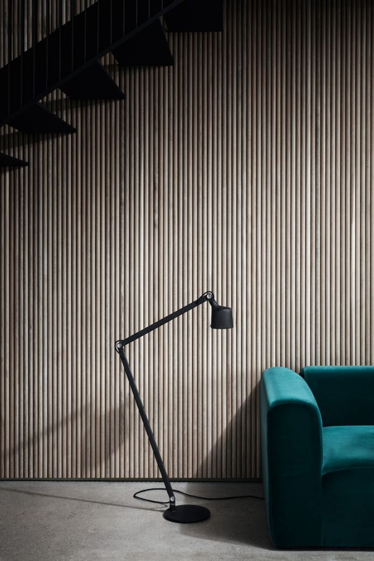 Schwarze Vipp Boden Leselampe neben einem grünen Sofa vor einer Wand mit Holzstäben und Treppe.