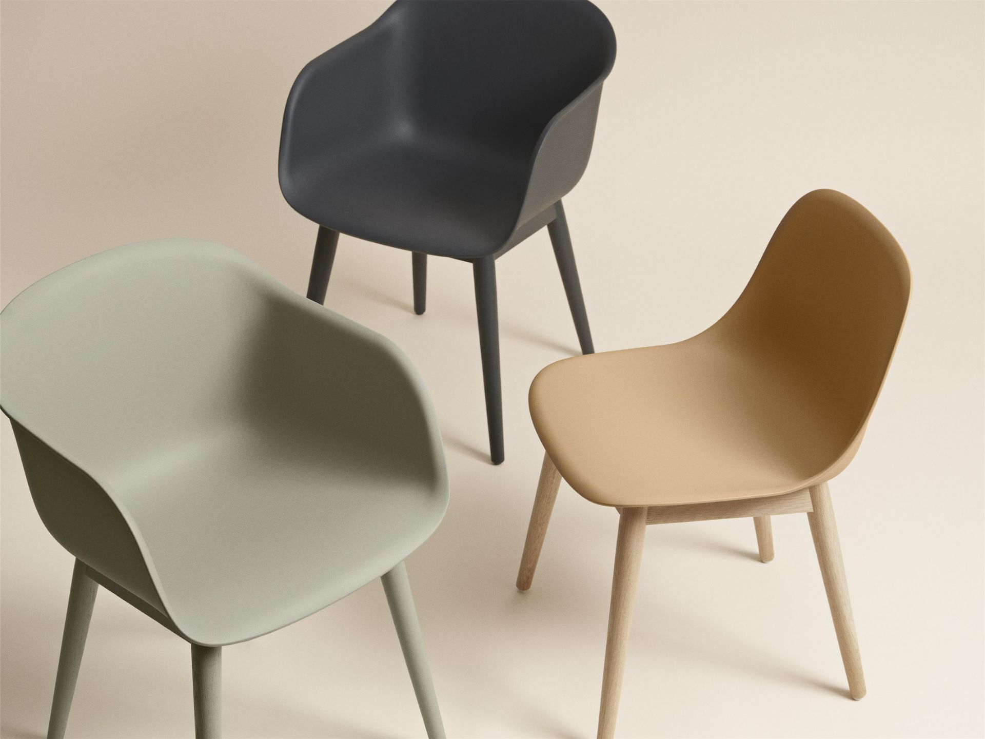 Drei Muuto Fiber Armchair Stühle mit Holzgestell in Schwarz, Beige und Grün auf beigem Hintergrund.