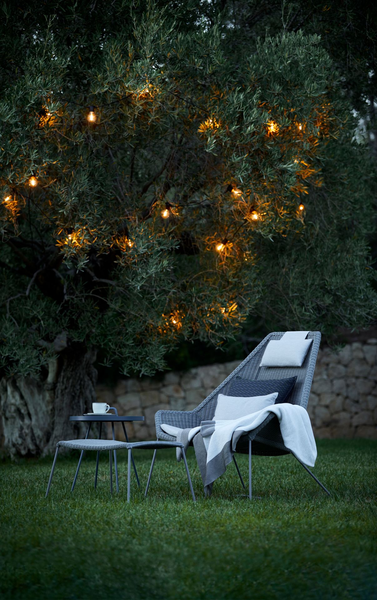 Breeze Highbacksessel von Cane-Line im Garten mit Beistelltisch und Lichterkette im Baum.