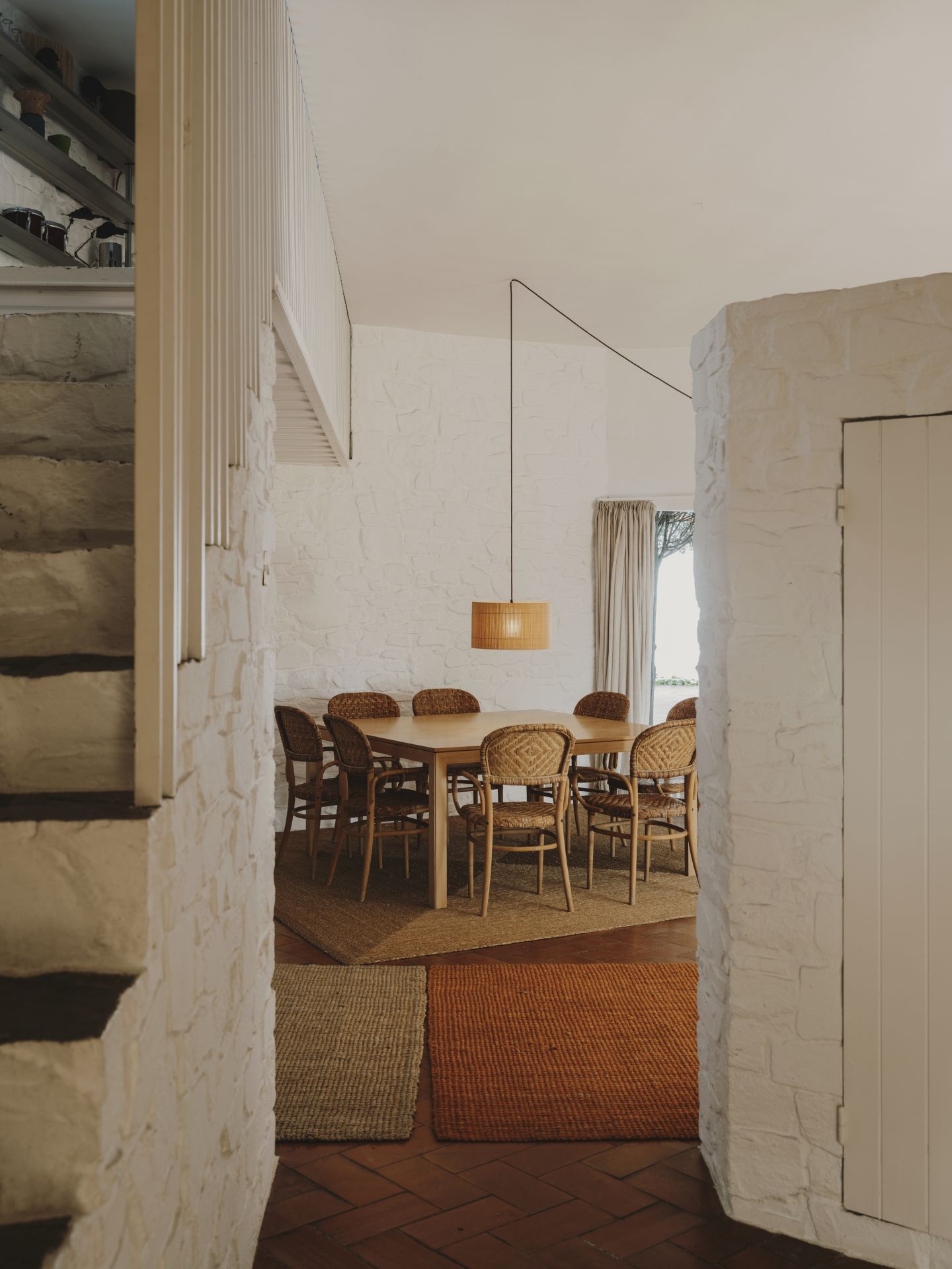 Esszimmer mit Holztisch, Rattan-Stühlen und gewebter Pendelleuchte in einem Haus mit weißen Ziegelwänden.