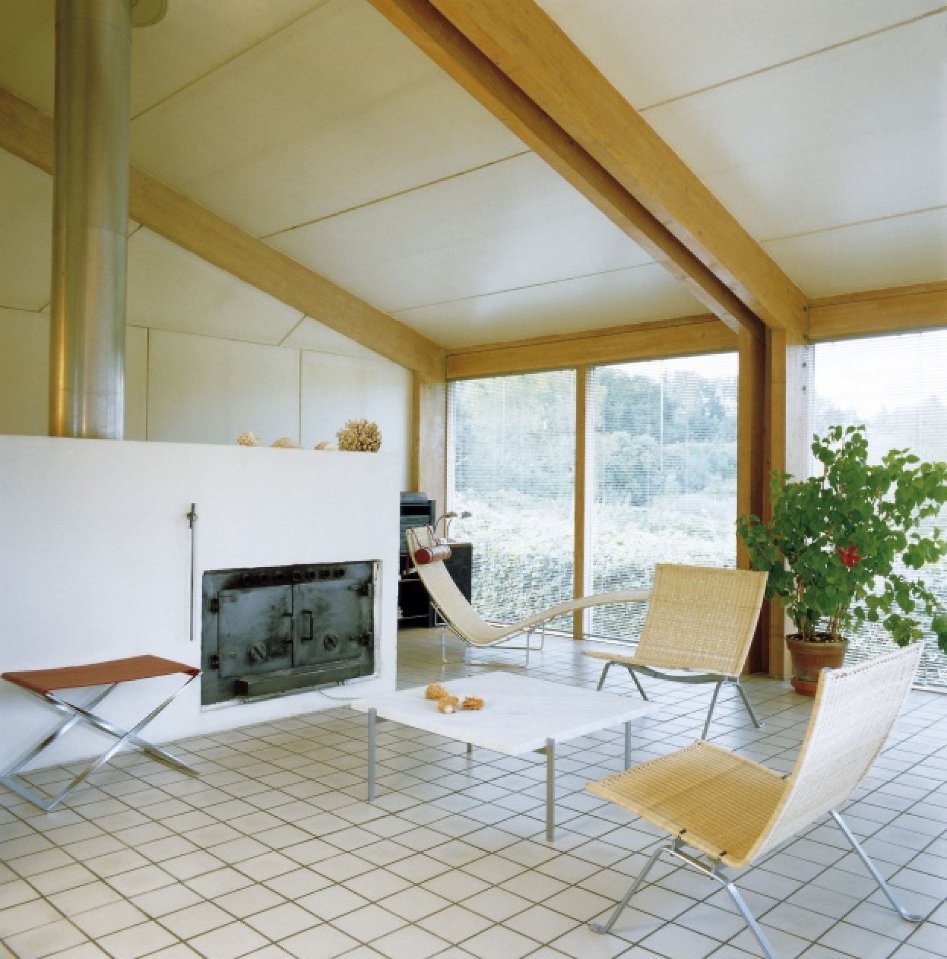 Heller Wohnraum mit PK 61 Couchtisch von Fritz Hansen, Stühlen, Liege und großem Fenster mit Blick ins Grüne.